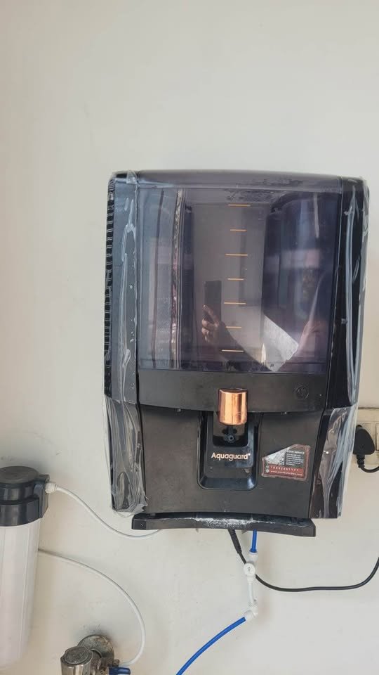 Aquaguard Enrich Aura 2X RO wall mounted installation showing inlet outlet water pipes connected to external pre filter and power plug on kitchen wall