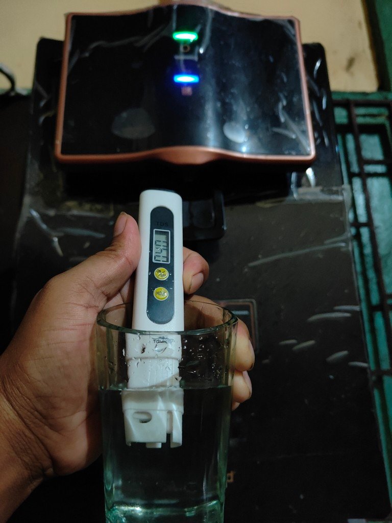 TDS meter dipped in a glass of purified RO water, with the digital display showing the TDS reading in ppm in front of a water purifier.